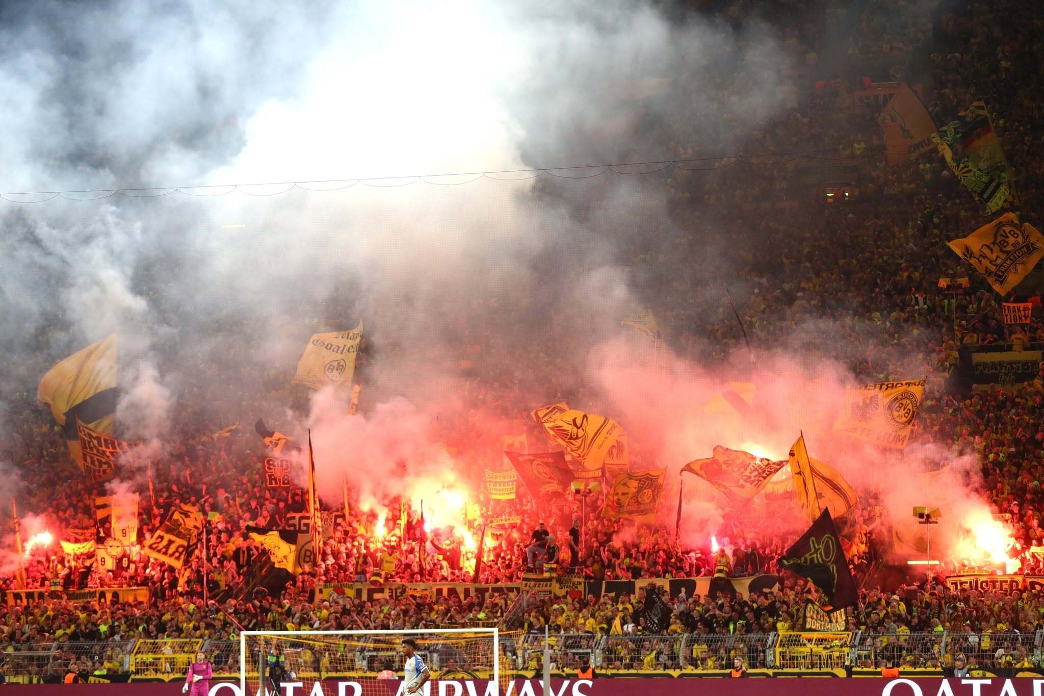 Eine große Masse an Fans. Am linken und rechten Bildrand halten einzelne Menschen bengalische Fackeln in die Höhe. Dazwischen zahlreiche schwarzgelbe Fahnen.