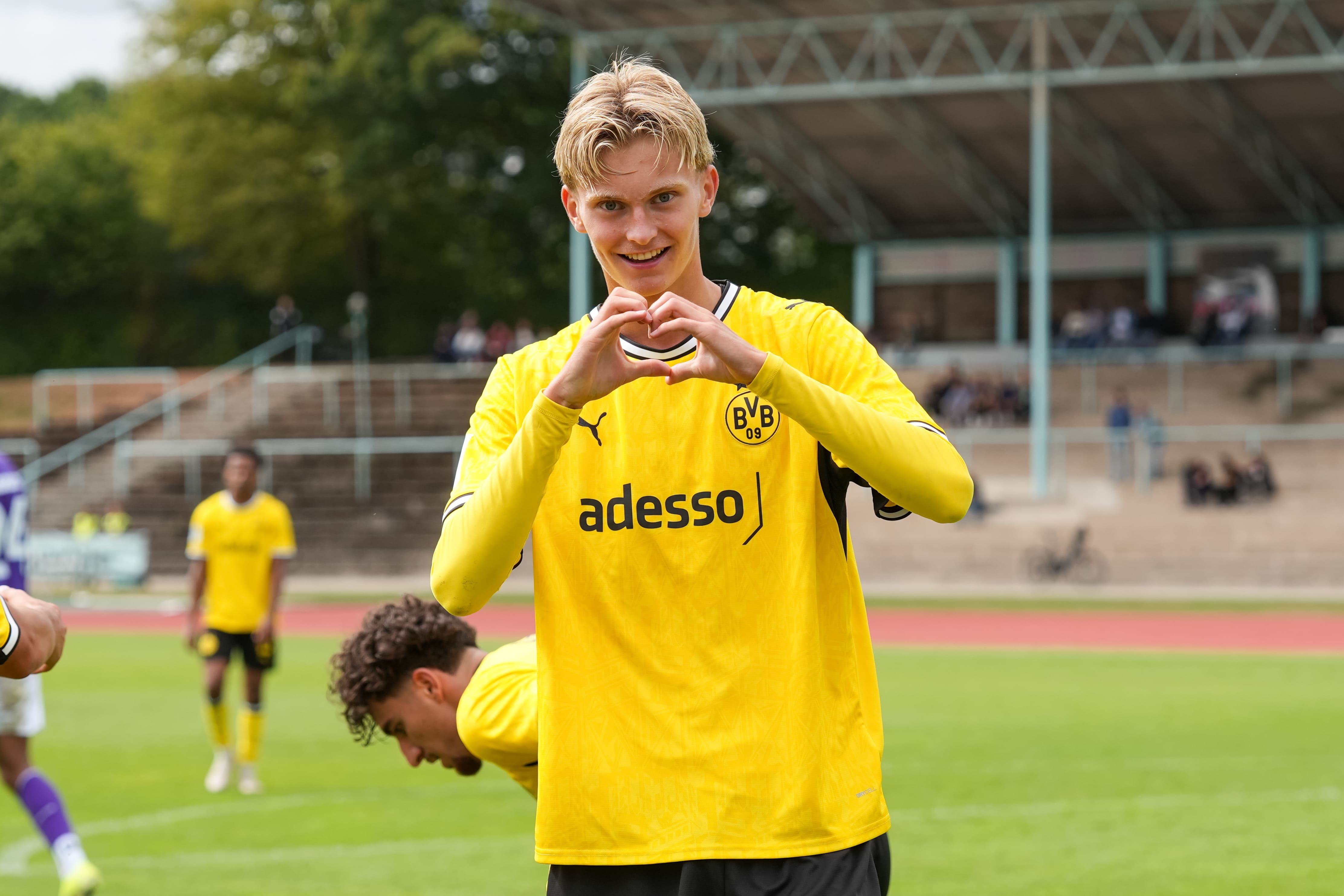 Ein junger blonder Spieler zeigt ein Herz in die Kamera. Hinter ihm beugt sich sein Mitspieler gerade herunter.