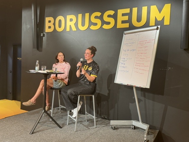 Josefine Schnieders und Merle Berg sitzen gemeinsam auf dem Podium. Rechts steht ein Flipchart.